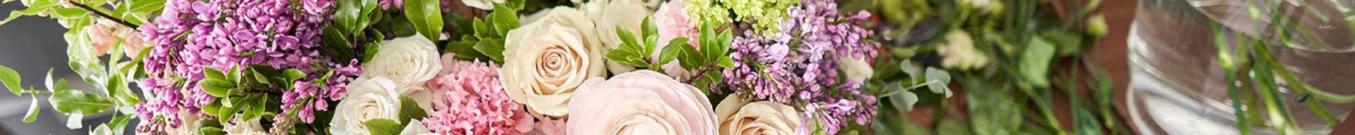 En person holder en kurv fyldt med farverige blomster, herunder roser, ranunkler og liljekonvaller. Der er også flere friske blomster på bordet, og stemningen er lys og indbydende. 